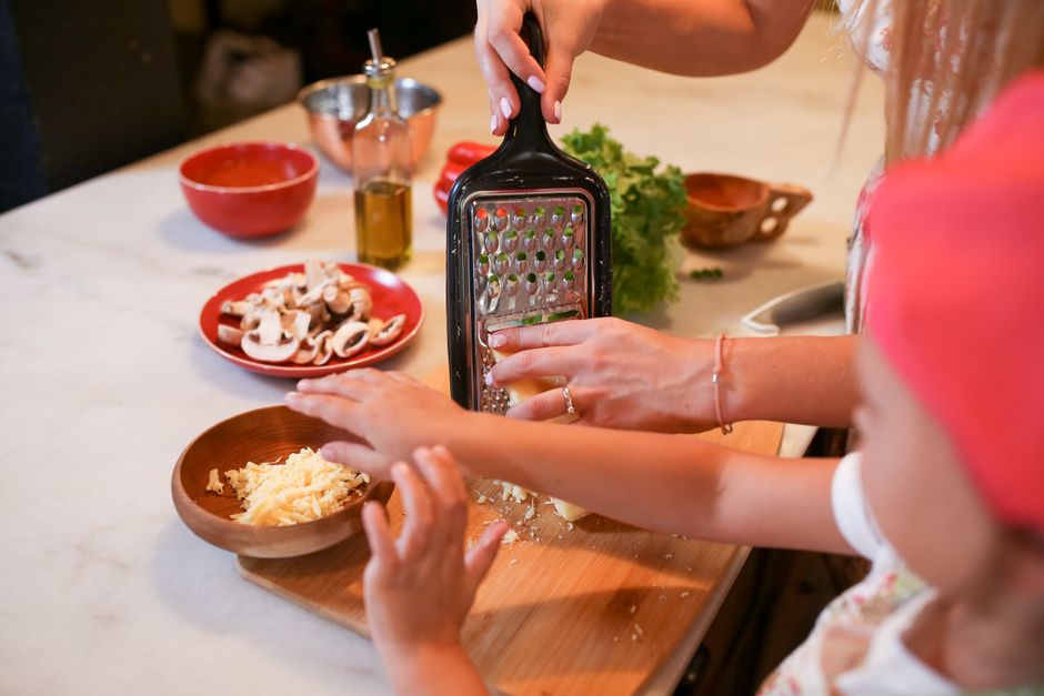 A mother and daughter grating cheese together on a kitchen counter with ingredients.